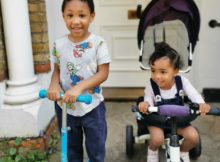 A preschool aged black boy on his scooter standing outside the door to his home next to his toddler sister on her trike smiling and looking happy